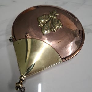 Könnte beinhalten: Ein kreisförmiger Bettwärmer aus Kupfer mit einem polierten Messing-Fleur-de-Lis-Emblem in der Mitte. Der Gegenstand verfügt über einen dreieckigen Messingaufsatz an der Basis, der mit einem kleinen, abgerundeten Griffteil mit einer Öse am Ende verbunden ist.