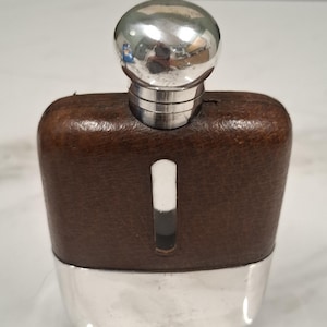 Pode incluir: Uma garrafa de bolso com a parte superior revestida a couro castanho com uma janela de visualização vertical em vidro, uma base em metal prateado polido e uma tampa de rosca arredondada em metal prateado com acabamento liso e refletor.