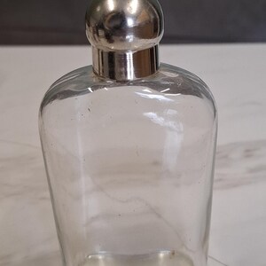Puede incluir: Una petaca de cristal transparente con cuerpo redondeado y un tapón de metal plateado liso y pulido. El recipiente presenta una base ancha y plana y un cuello estrecho, con un diseño sencillo y transparente que permite ver el interior.