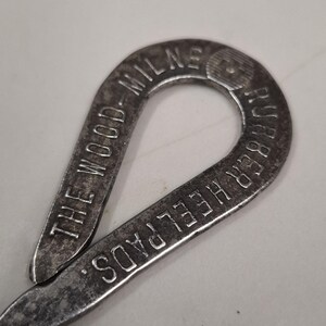 May include: Close-up of a vintage metal shoe repair tool. The tool is a loop shape with the words "THE WOOD - MILNE RUBBER HEELPADS" embossed on the surface. The metal has a worn, aged appearance.