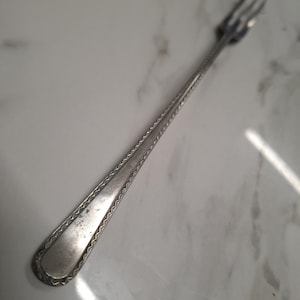 Peut inclure: Une fourchette en argent avec trois dents et un long manche décoratif. Le manche présente un motif en zigzag détaillé sur les bords. La fourchette est probablement vintage ou antique, montrant des signes d'usure.
