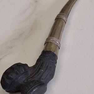 Puede incluir: Una pipa de fumar larga y curva con una base tallada en negro mate oscuro con patrones texturizados. El tallo está hecho de un material metálico plateado liso, acentuado por dos bandas decorativas de intrincado alambre de plata trenzado.