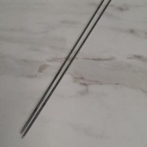 May include: A pair of thin, straight, metallic grey knitting needles. These long, slender tools feature a smooth, uniform cylindrical shape with slightly tapered, rounded points at one end, designed for manual textile crafting and yarn work.