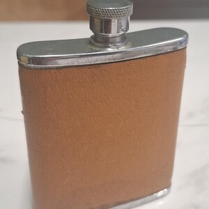 May include: A rectangular metal flask with a curved body wrapped in a tan textured material. The flask features a polished silver-tone metal top, a threaded neck, and a matching silver-tone screw-on cap with a knurled edge for a secure grip.