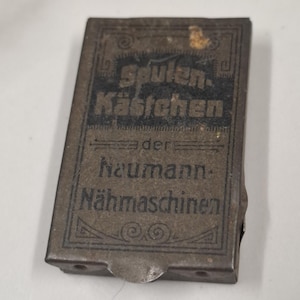 May include: A vintage metal spool box with the text "Spulen Kästchen der Naumann Nähmaschinen" in black lettering. The box is rectangular with a hinged lid and a small metal clasp. The box has a worn, antique appearance.