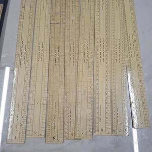 May include: A collection of vintage wooden rulers, each with inch and foot markings. The rulers are beige with black markings and text, arranged on a light-colored surface. The rulers are of varying lengths, with some showing signs of age.