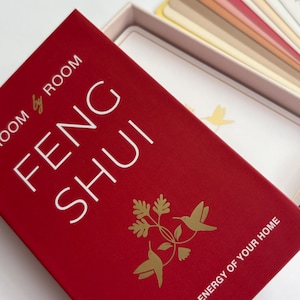 Op de afbeelding: Een rode doos met de woorden "FENG SHUI" in het wit. De doos heeft gouden kolibrie-illustraties en de tekst "ENHANCE THE ENERGY OF YOUR HOME". Binnenin is een kaartspel zichtbaar.