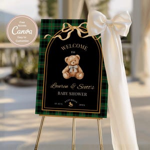 May include: A baby shower welcome sign on a gold easel. The sign has a black background with a teddy bear illustration and gold accents. The sign reads "Welcome to Lauren & Scott's Baby Shower". A white bow and ribbon are attached to the easel.