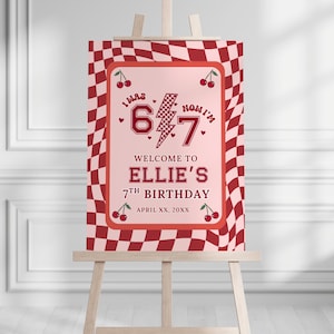 May include: A birthday party sign on an easel. The sign has a pink background with a red checkered border. The text reads "I was 6 now I'm 7" with "Welcome to Ellie's 7th Birthday" and the date.