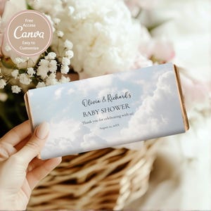 Op de afbeelding: Een baby shower gunst met een hemel-thema ontwerp. De verpakking heeft een blauwe lucht met pluizige witte wolken en de tekst "Olivia & Richard's BABY SHOWER". De gunst wordt in een hand gehouden, met bloemen op de achtergrond.