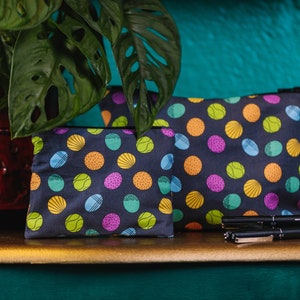 May include: Two dark blue zippered pouches with a colourful dot pattern. The dots are in various colours and designs, including yellow, green, orange, and purple. The pouches are on a wooden surface with black pens.