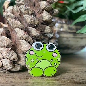 May include: A green frog enamel pin with black eyes, a white smile, and pink cheeks. The frog has a spotted pattern and is sitting with its legs crossed.