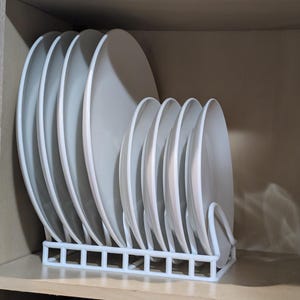 Puede incluir: Un estante blanco de plástico para platos que sostiene una pila de platos blancos dentro de un armario de madera. Los platos están dispuestos verticalmente, mostrando su diseño limpio y minimalista. El estante proporciona un almacenamiento organizado para la vajilla.