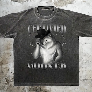 Puede incluir: Camiseta gris oscuro con un lobo con sombrero de vaquero. El texto "CERTIFIED GOONER" está impreso encima y debajo de la ilustración. La camiseta tiene un aspecto vintage y desgastado.