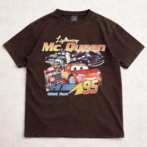 Puede incluir: Camiseta marr&oacute;n con un dise&ntilde;o de Rayo McQueen y otros personajes de Cars. El dise&ntilde;o incluye el texto "Lightning Mc.Queen" y "95". La camiseta tiene un aspecto vintage y desgastado.