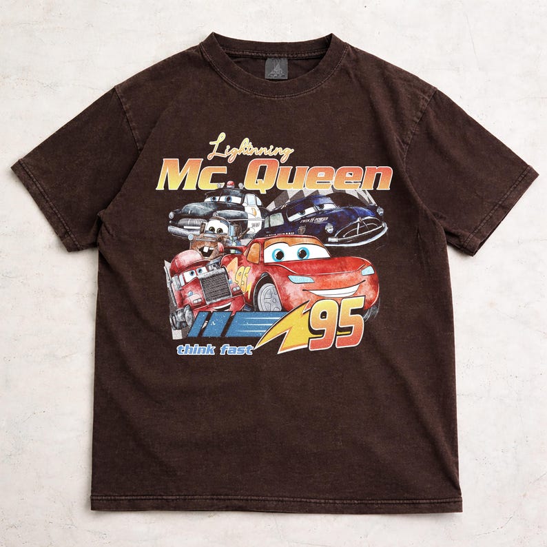Puede incluir: Camiseta marr&oacute;n con un dise&ntilde;o de Rayo McQueen y otros personajes de Cars. El dise&ntilde;o incluye el texto "Lightning Mc.Queen" y "95". La camiseta tiene un aspecto vintage y desgastado.