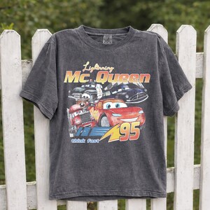 Puede incluir: Camiseta gris oscuro con Lightning McQueen de la pel&iacute;cula Cars. El gr&aacute;fico incluye el texto "Lightning Mc. Queen", "95" y "think fast", junto con im&aacute;genes del personaje y otros veh&iacute;culos.