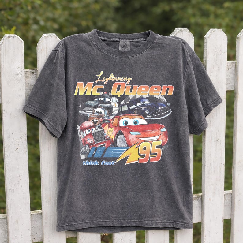 Puede incluir: Camiseta gris oscuro con Lightning McQueen de la pel&iacute;cula Cars. El gr&aacute;fico incluye el texto "Lightning Mc. Queen", "95" y "think fast", junto con im&aacute;genes del personaje y otros veh&iacute;culos.