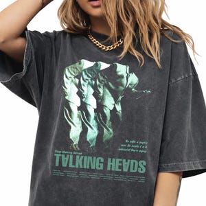 May include: Dark gray oversized t-shirt featuring a green and white graphic of three figures. The shirt has the text "TALKING HEADS" in green, with additional text above and below the band's name. The shirt has a vintage, worn look.