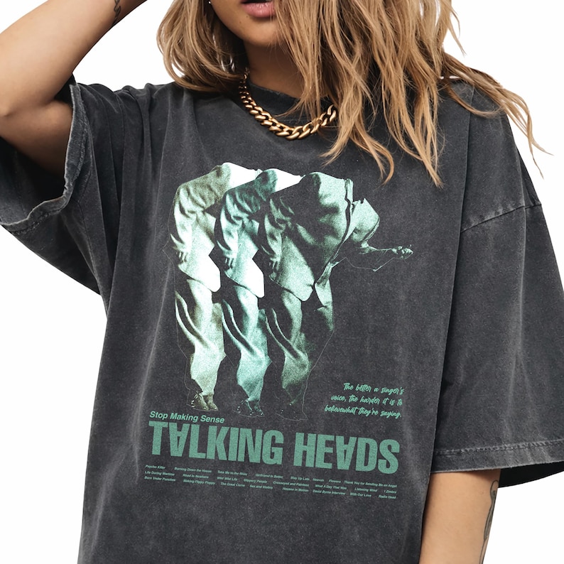 May include: Dark gray oversized t-shirt featuring a green and white graphic of three figures. The shirt has the text "TALKING HEADS" in green, with additional text above and below the band's name. The shirt has a vintage, worn look.