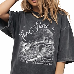 Puede incluir: Camiseta gris oscuro con la inscripción "The Shire" y una ilustración de una puerta de hobbit. La cita dice: "Todo lo que tenemos que decidir es qué hacer con el tiempo que se nos da."
