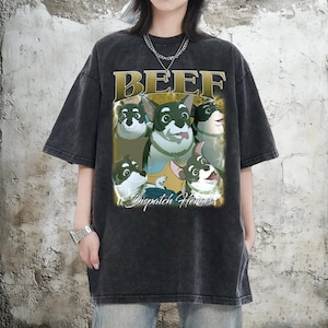 Puede incluir: Camiseta gris oscuro con un gráfico de perros de dibujos animados y el texto "BEEF" en oro arriba y "Dispatch Heroes" abajo. Los perros tienen collares verdes. La camiseta tiene mangas cortas y un corte holgado.
