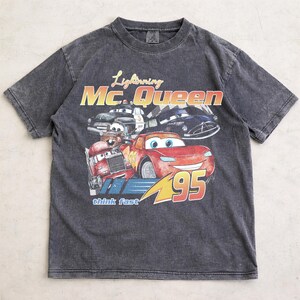 Puede incluir: Camiseta gris carb&oacute;n con un dise&ntilde;o de Rayo McQueen y otros personajes de la pel&iacute;cula Cars. El dise&ntilde;o incluye el texto "Lightning Mc. Queen" y "95". La camiseta tiene un aspecto vintage y desgastado.