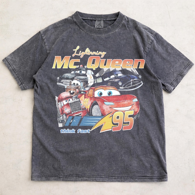 Puede incluir: Camiseta gris carb&oacute;n con un dise&ntilde;o de Rayo McQueen y otros personajes de la pel&iacute;cula Cars. El dise&ntilde;o incluye el texto "Lightning Mc. Queen" y "95". La camiseta tiene un aspecto vintage y desgastado.