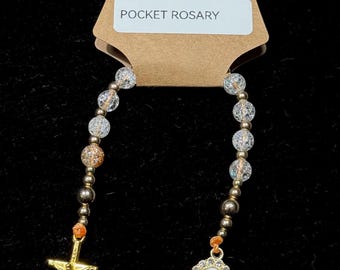 May include: Pocket Rosary featuring clear and gold beads, a gold crucifix, and a gold and white Virgin Mary pendant. The rosary is attached to a brown tag with the text "POCKET ROSARY".