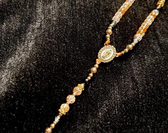 May include: A gold-coloured rosary with a detailed cross pendant and an oval centrepiece featuring an image. The beads are a mix of gold, clear, and iridescent colours, strung on a delicate chain. The rosary is displayed against a dark, textured background.