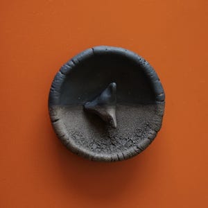 Könnte beinhalten: Eine kleine, runde, dunkelgraue Keramikschale mit erhöhtem Rand und strukturierter Oberfläche. Ein dunkelgraues, dreieckiges Objekt befindet sich in der Mitte. Die Schale steht vor einem orangefarbenen Hintergrund.