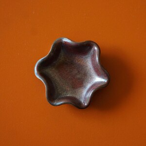 Könnte beinhalten: Eine kleine, sternförmige Keramikschale mit metallischer, gesprenkelter Oberfläche in Braun- und Bronzetönen. Die Schale hat einen welligen, unregelmäßigen Rand und liegt auf einer orangefarbenen Oberfläche. Die Schale ist leer.