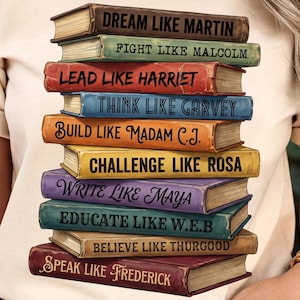 May include: Beige t-shirt featuring a stack of books with inspirational phrases. Each book has a different color and text, including "DREAM LIKE MARTIN" and "SPEAK LIKE FREDERICK". The design is a tribute to influential figures.