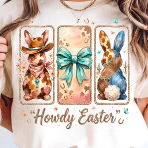 Puede incluir: Camiseta color crema con tres ilustraciones enmarcadas de conejos de Pascua. Los conejos están adornados con sombreros de vaquero, lazos turquesa y diseños de patchwork. El texto "Howdy Easter" está escrito en una fuente de guion debajo de las ilustraciones.