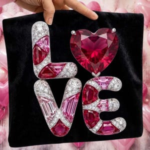 Puede incluir: Un fondo de terciopelo negro muestra la palabra "LOVE" escrita con letras enjoyadas. Las letras "L", "V" y "E" est&aacute;n incrustadas con diamantes y presentan piedras preciosas rosas y rojas. Un gran rub&iacute; en forma de coraz&oacute;n se encuentra encima de la "E".