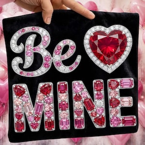 Puede incluir: Coj&iacute;n de terciopelo negro con las palabras "Be Mine" escritas con brillantes piedras preciosas rojas, rosas y transparentes. Tambi&eacute;n presenta una gran gema en forma de coraz&oacute;n. El coj&iacute;n est&aacute; sostenido por una mano.