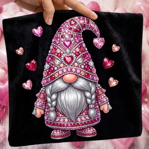 Puede incluir: Un coj&iacute;n negro con un dise&ntilde;o de gnomo. El gnomo tiene una larga barba blanca, una nariz rosada y un sombrero adornado con corazones y joyas. Gemas rosas en forma de coraz&oacute;n est&aacute;n dispersas alrededor del gnomo. El atuendo del gnomo est&aacute; decorado con joyas rosas y rojas.