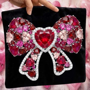 Puede incluir: Un broche grande con forma de lazo enjoyado con un coraz&oacute;n en el centro. El lazo est&aacute; hecho de varias tonalidades de piedras preciosas rojas, rosas y transparentes engastadas en un metal plateado. El broche se muestra sobre una superficie negra.
