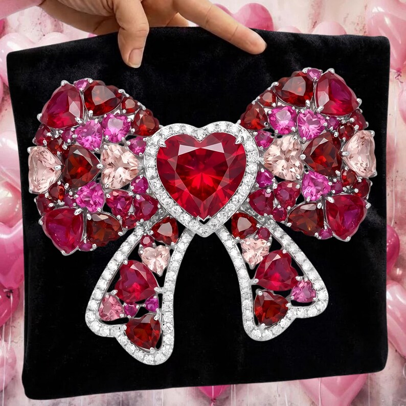 Puede incluir: Un broche grande con forma de lazo enjoyado con un coraz&oacute;n en el centro. El lazo est&aacute; hecho de varias tonalidades de piedras preciosas rojas, rosas y transparentes engastadas en un metal plateado. El broche se muestra sobre una superficie negra.