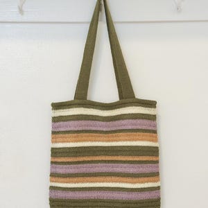 May include: A handmade crochet tote bag with olive green handles and body. The bag features horizontal stripes in shades of olive green, cream, lavender, and peach. The bag is hanging against a white wall.