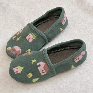 May include: A pair of olive green knit slippers featuring a repeating pattern of small pink houses and light yellow trees. The slippers have a rounded toe, a low-cut opening, and a dark brown interior lining, providing a simple and functional design.