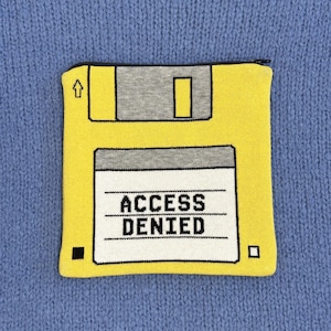 Pode incluir: Bolsa amarela em forma de disquete com a inscrição preta "ACCESS DENIED". A bolsa tem um zíper preto e está sobre um fundo azul tricotado. O design imita um disquete clássico de 3,5 polegadas.