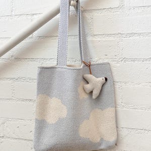 May include: A light blue tote bag with a cloud pattern and a white bird charm. The bag has a woven texture and a light-colored interior. The bag is hanging from a white wooden bar against a white brick wall.