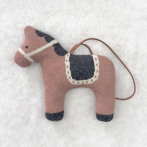 May include: A plush horse ornament in shades of dusty rose, gray, and white. The horse has a saddle, bridle, and a dark gray mane. A brown leather loop is attached for hanging. The horse is approximately 5 inches long.