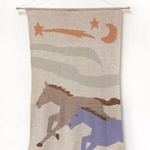 May include: A rectangular knit wall hanging suspended from a wooden rod, featuring a beige background with a brown horse and a periwinkle horse running. Above them are two sage green clouds, an orange shooting star, and an orange crescent moon.