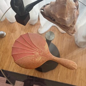 May include: A selection of decorative items on a glass table. Features a bronze sculpture, a large seashell, and a fan-shaped object with a red and yellow dotted pattern.