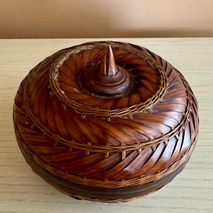 May include: A round, woven basket with a spiral design on the lid. The basket is made of brown bamboo and has a conical knob at the centre. The basket's sides are a darker brown, and the overall shape is reminiscent of a nautilus shell.