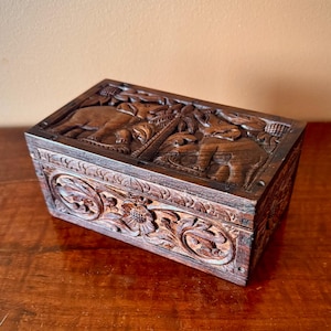 May include: A rectangular wooden box with intricate carvings. The dark brown box features detailed relief carvings of elephants and floral patterns. The lid and sides are adorned with ornate designs, showcasing a handcrafted aesthetic.