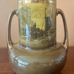 Può includere: Vaso vintage in ceramica con due manici e base larga. Il vaso presenta una scena paesaggistica con una barca e la scritta "ST. HELENS". Il vaso ha una finitura lucida con dettagli dorati sui manici e sul bordo.