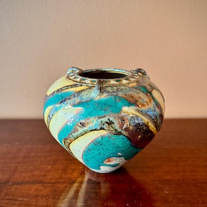 Può includere: Un vaso in ceramica con corpo arrotondato e ampia apertura. Il vaso presenta una vibrante tavolozza di colori verde acqua, giallo, marrone e blu. L'esterno ha una finitura strutturata e smaltata. Due piccole maniglie sono sul bordo.